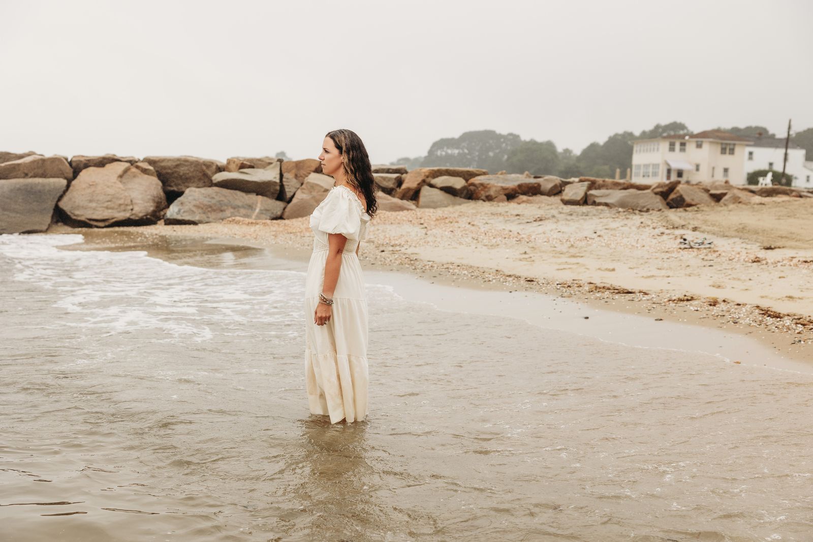 Ali Lacroix in a cream dress, standing in shallow water on a foggy Connecticut beach and looking out over the water.