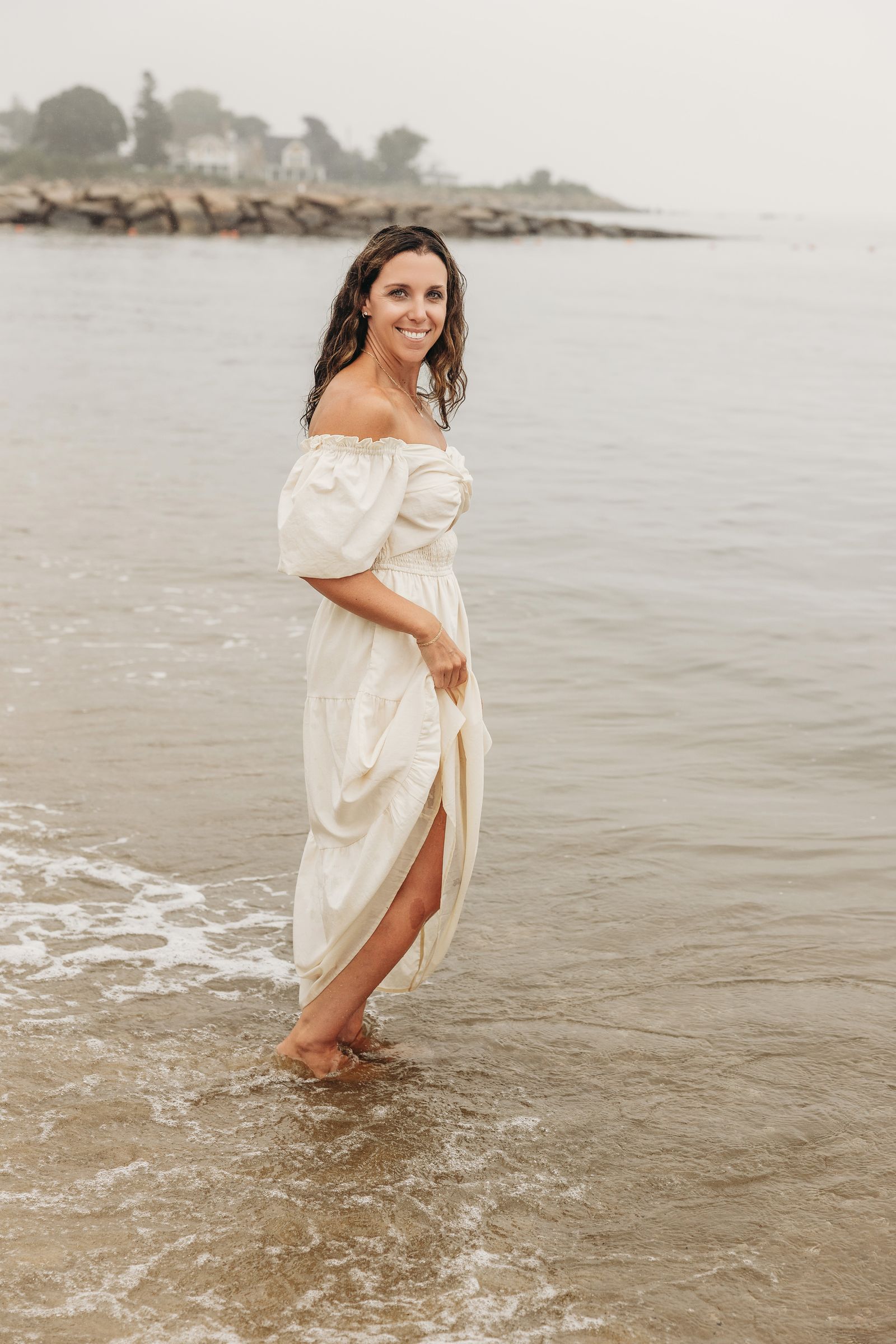 Ali Lacroix smiling at the camera, standing in shallow water on a foggy Connecticut beach in a cream dress.