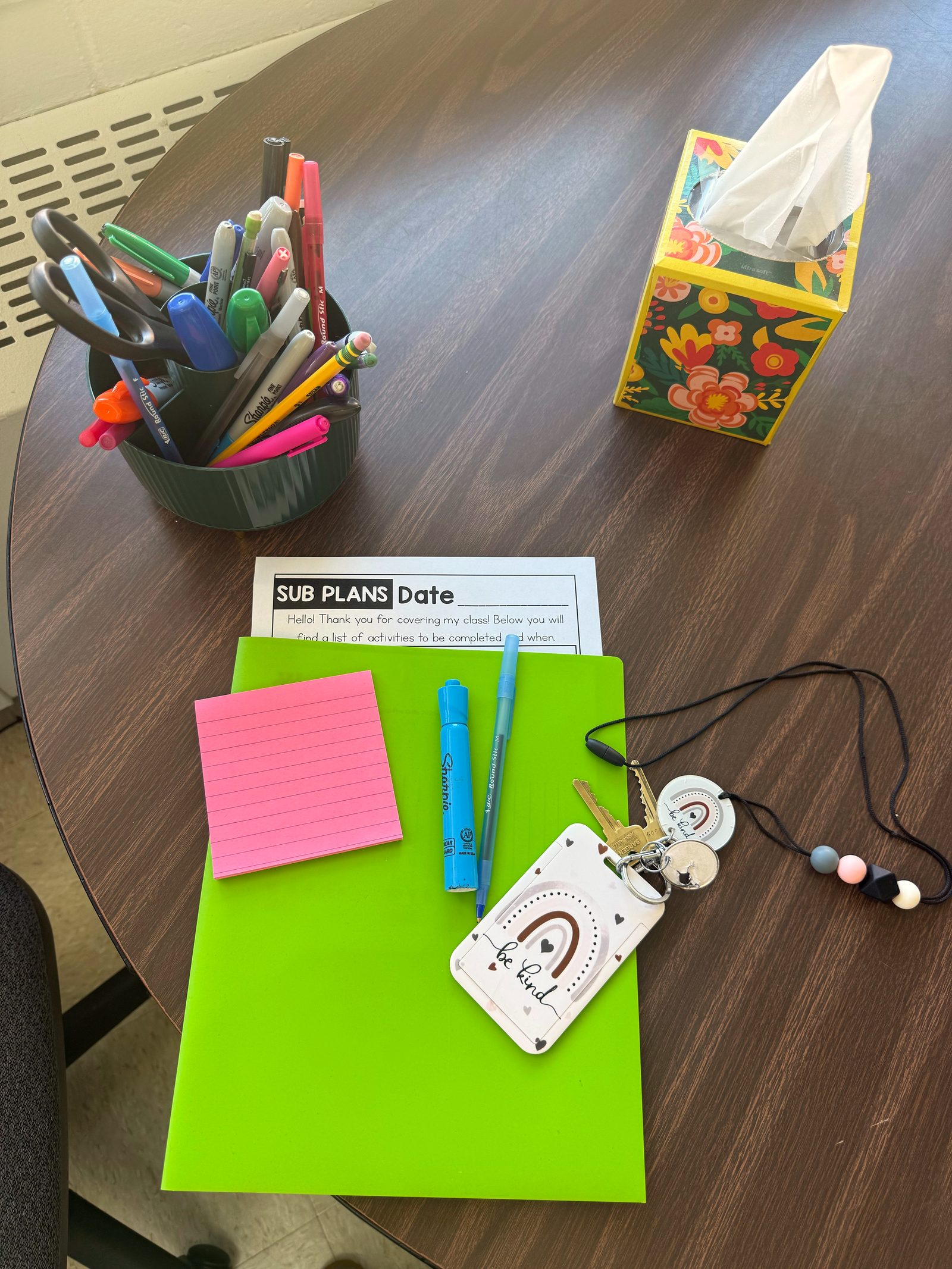 Overhead of Ali's desk with a sub plans sheet, a green folder, pink sticky notes, a highlighter, and a be kind lanyard.